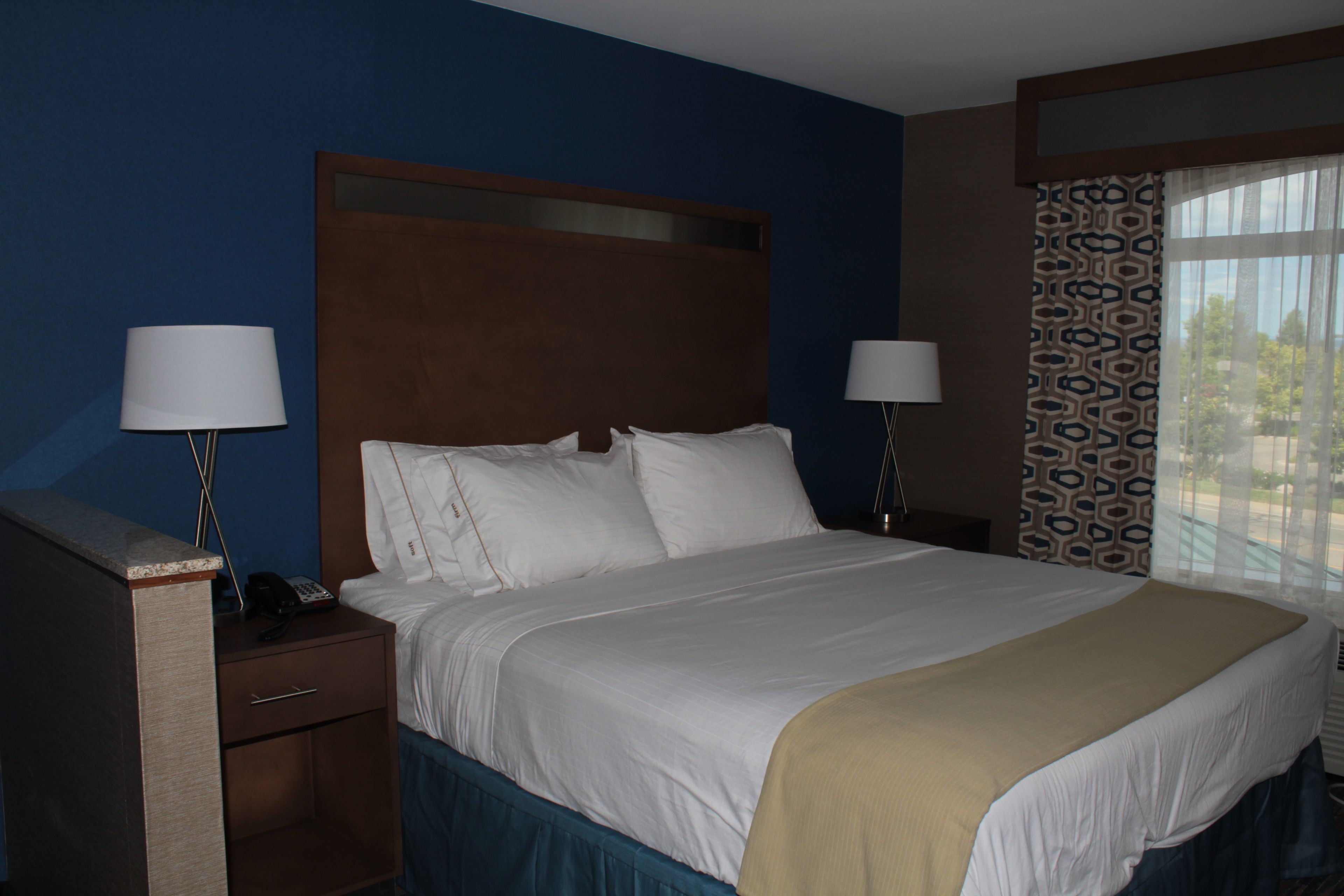 A hotel room with a large bed, two bedside tables with lamps, a wooden headboard, and a window with patterned curtains.