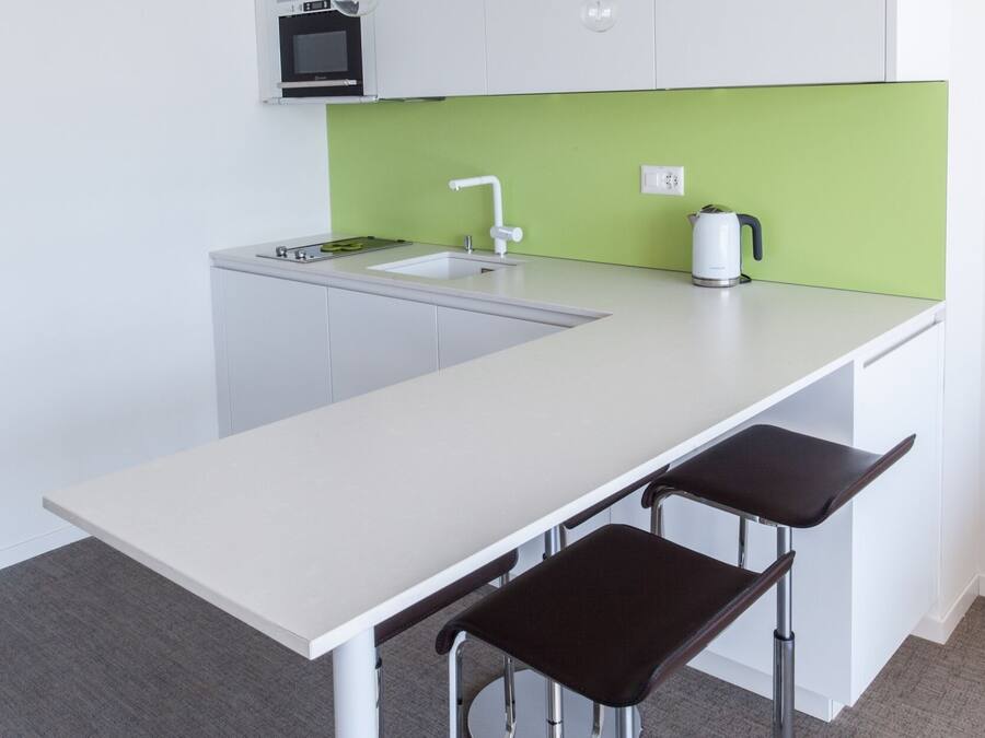 A modern kitchen with a white countertop, green backsplash, and a built-in oven. There are two bar stools and a white sink.