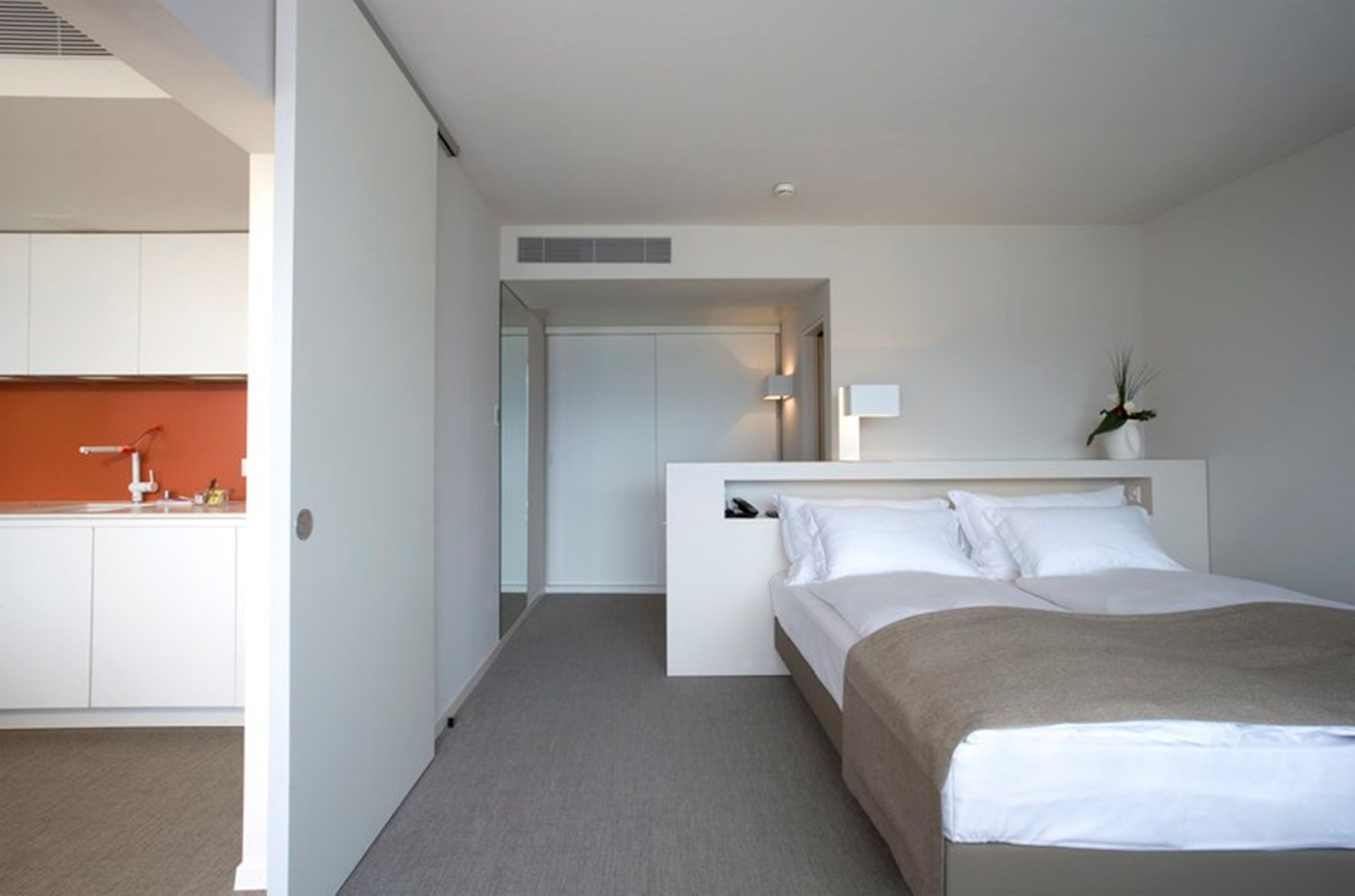Una habitación de hotel moderna con una cama, una mesita de noche, una lámpara y vistas a la cocina.
