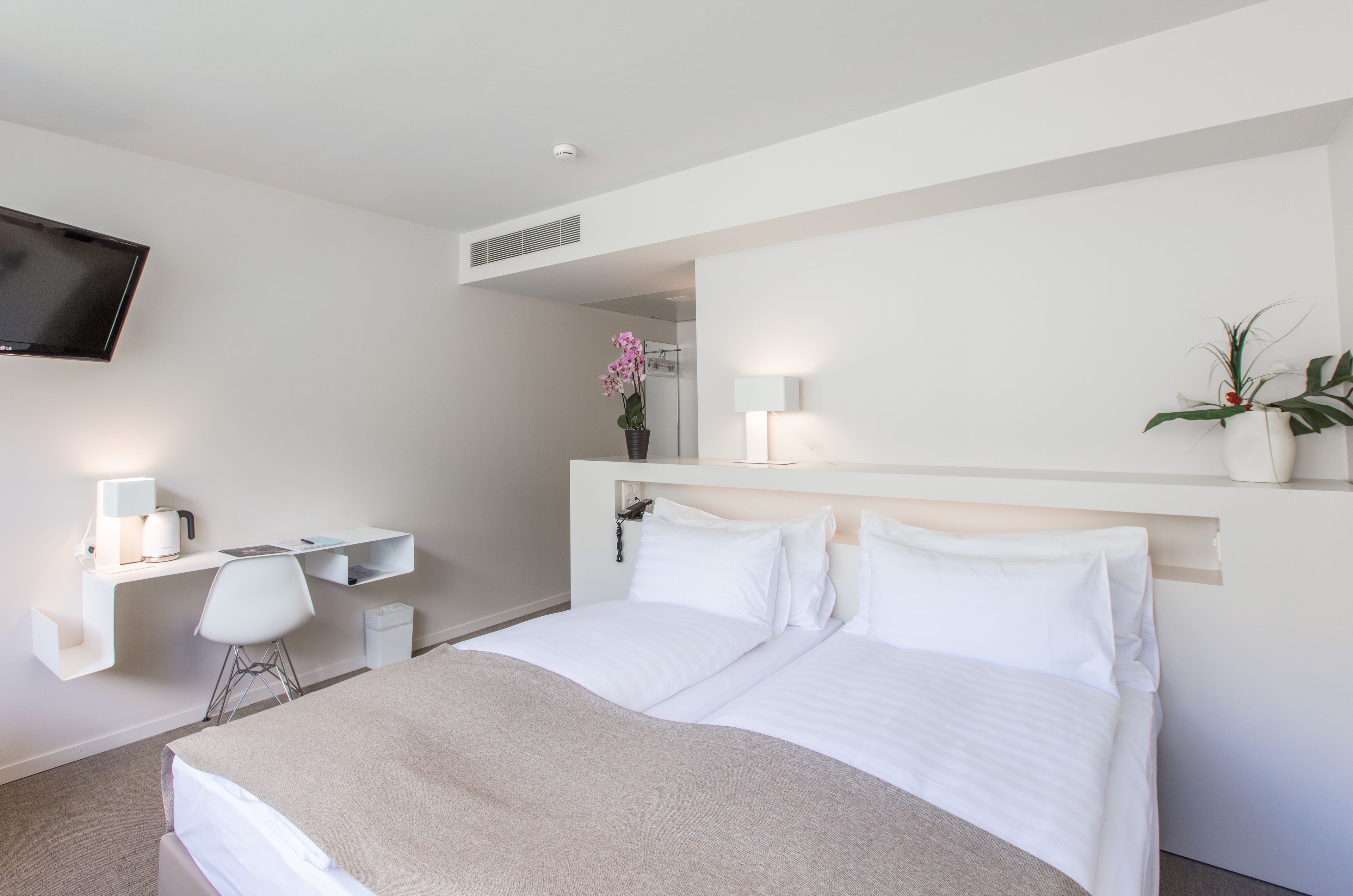 Una habitación de hotel moderna con una cama, un escritorio con silla, un televisor montado en la pared y una planta en maceta.