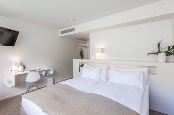 A modern hotel room with a bed, a desk with a chair, a TV mounted on the wall, and a potted plant.