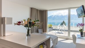 Habitación de hotel con una ventana grande, un escritorio, una silla, un televisor y vistas a las montañas y al mar.