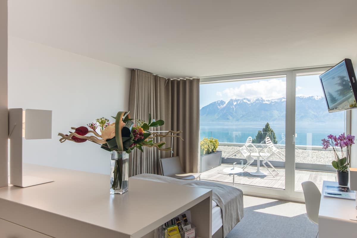 Hotelkamer met groot raam, bureau, stoel, televisie en uitzicht op bergen en zee.