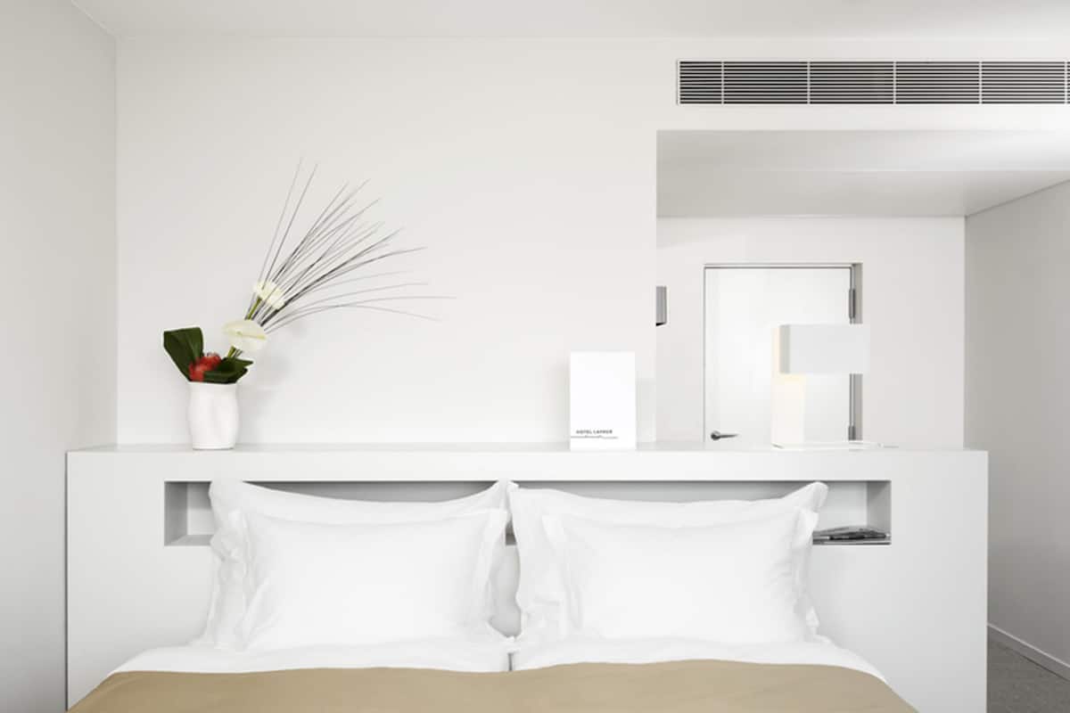 Een moderne hotelkamer met een wit bed, een nachtkastje met een bloevas, een spiegel en een luchtventilatie rooster aan de muur.