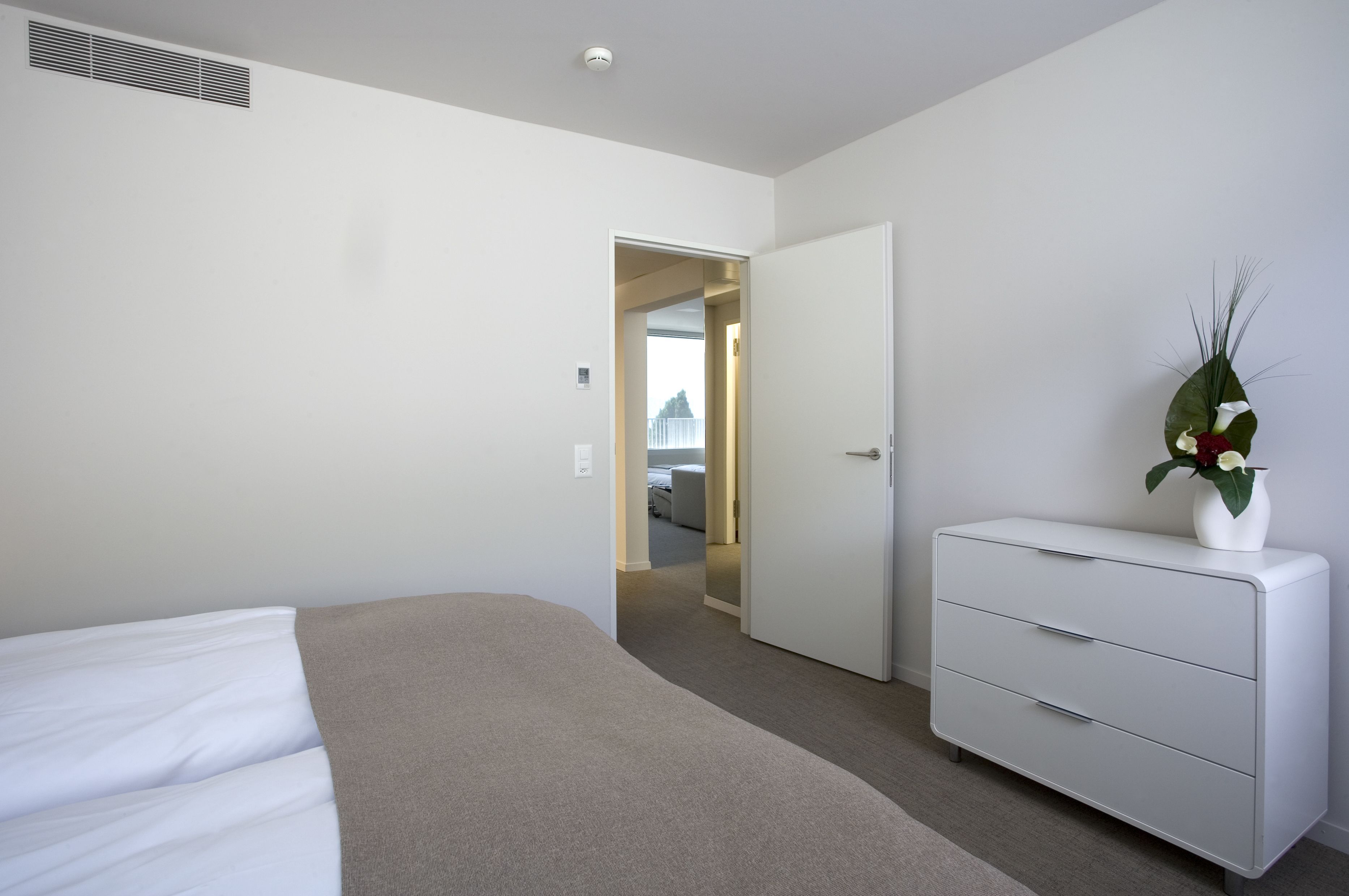 A modern hotel room with a bed, a dresser, and a door leading to another room.