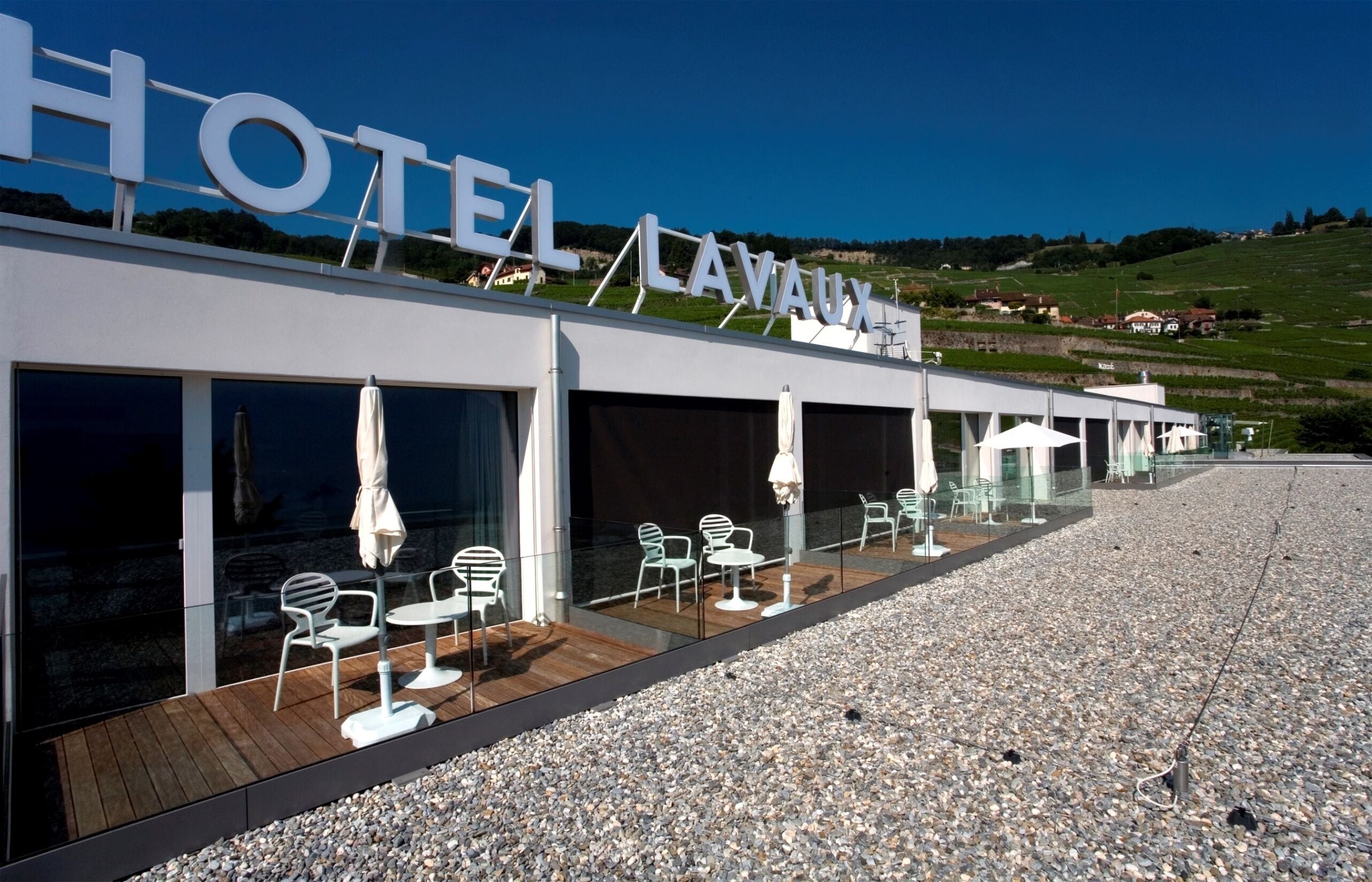 Een hotel met een modern design, met een opvallend bord, een terras met tafels en stoelen en uitzicht op het omliggende landschap.