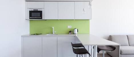 Una cocina moderna con armarios blancos, salpicadero verde y microondas integrado. Hay una encimera blanca con un fregadero y una tetera. Dos taburetes negros están ubicados en la barra.