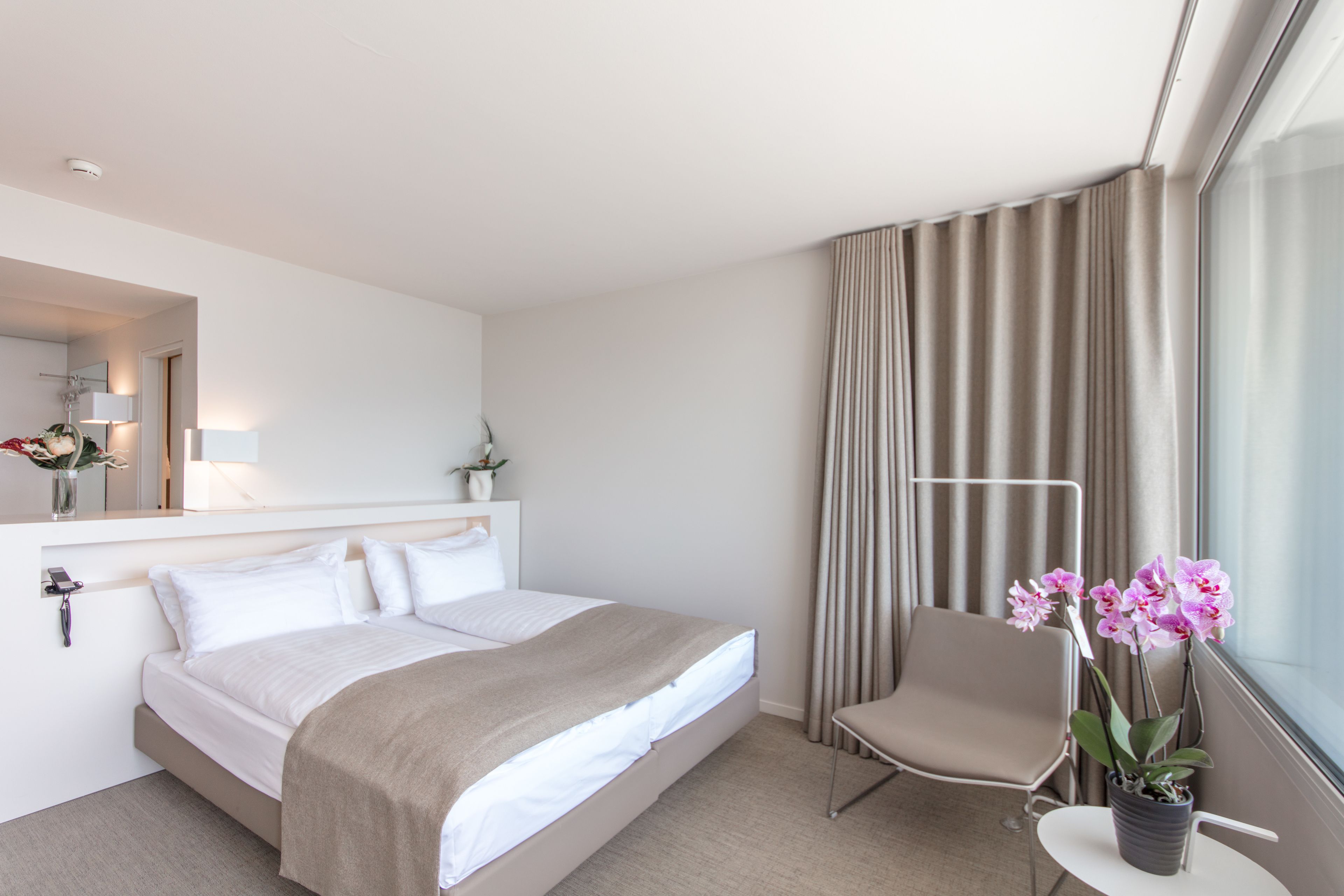 A hotel room with a bed, a chair, a small table with a plant, a window with curtains, and a mirror.