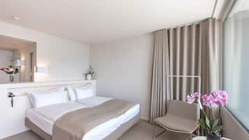 Ein Hotelzimmer mit Bett, Sessel, kleinem Tisch mit Pflanze, Fenster mit Vorhängen und Spiegel.
