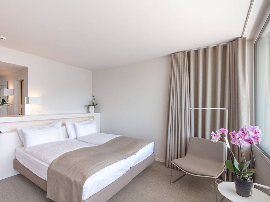 Een hotelkamer met een bed, een stoel, een klein tafeltje met een plant, een raam met gordijnen en een spiegel.