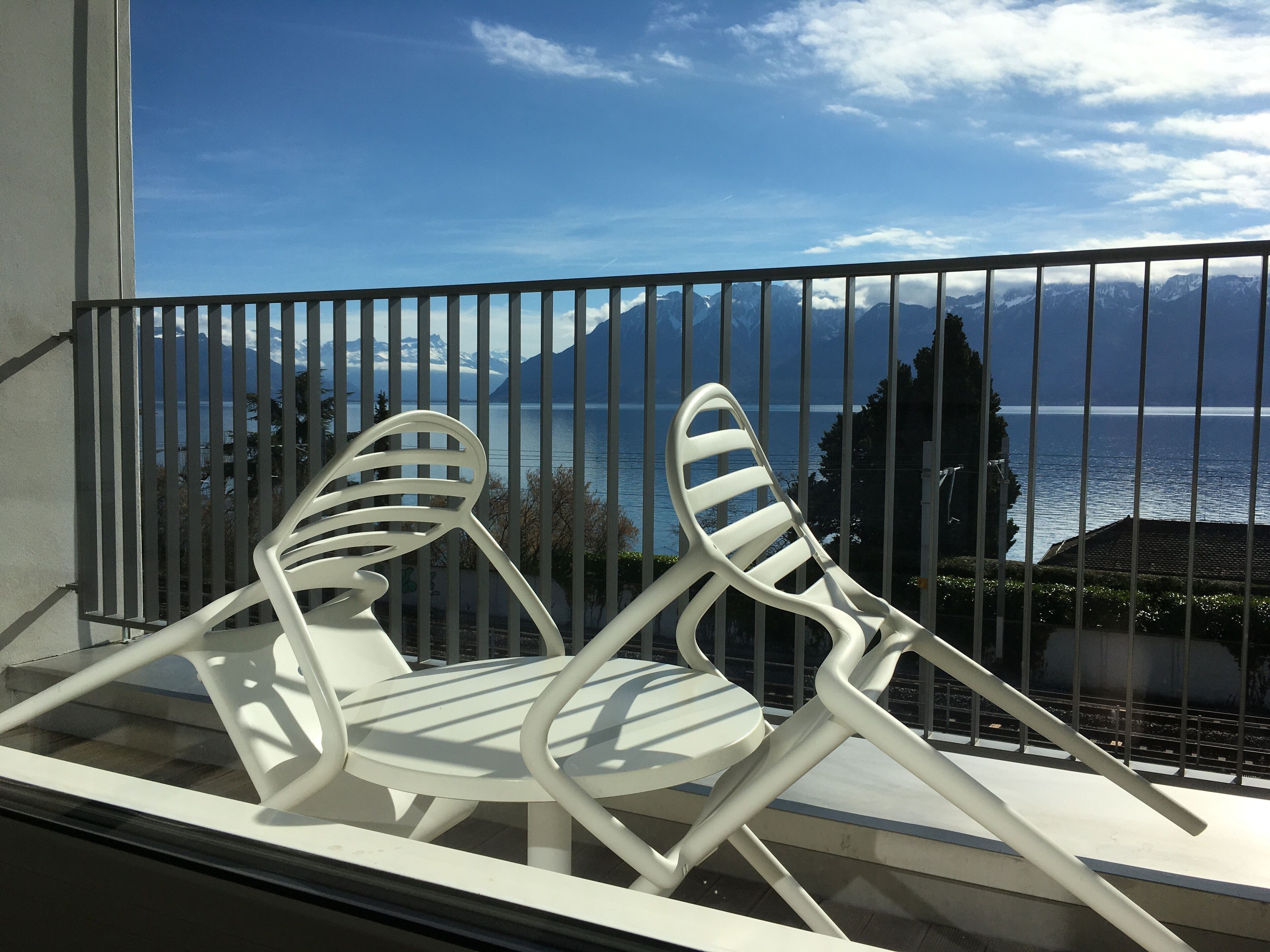 Un balcon avec deux chaises blanches et une table, d’où l’on peut admirer un plan d’eau et des montagnes.
