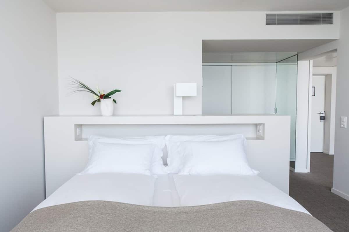 Een moderne hotelkamer met een groot bed, een nachtkastje, een kleine plant in pot en een douchecabine met glazen wanden.