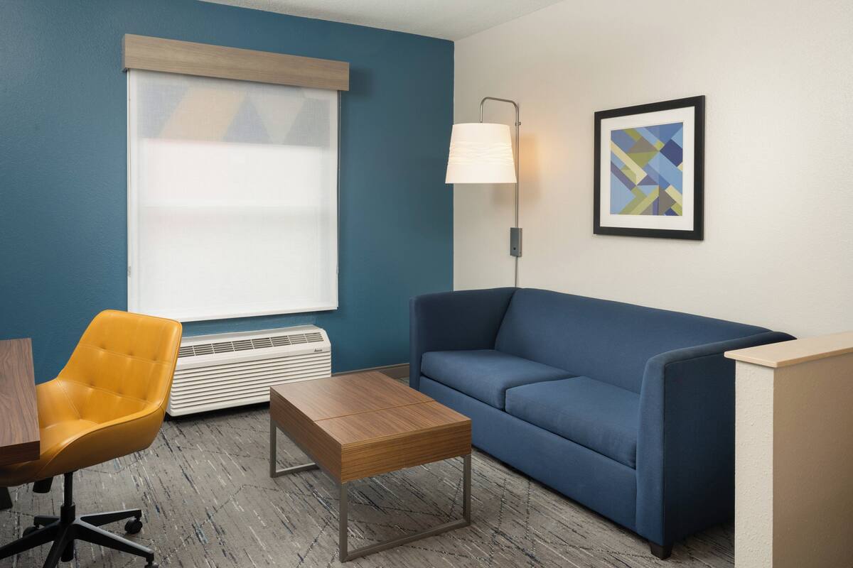 Een kamer met een blauwe wand, een blauwe bank, een gele stoel, een houten tafel, een vloerlamp en een lijst met een abstract schilderij.