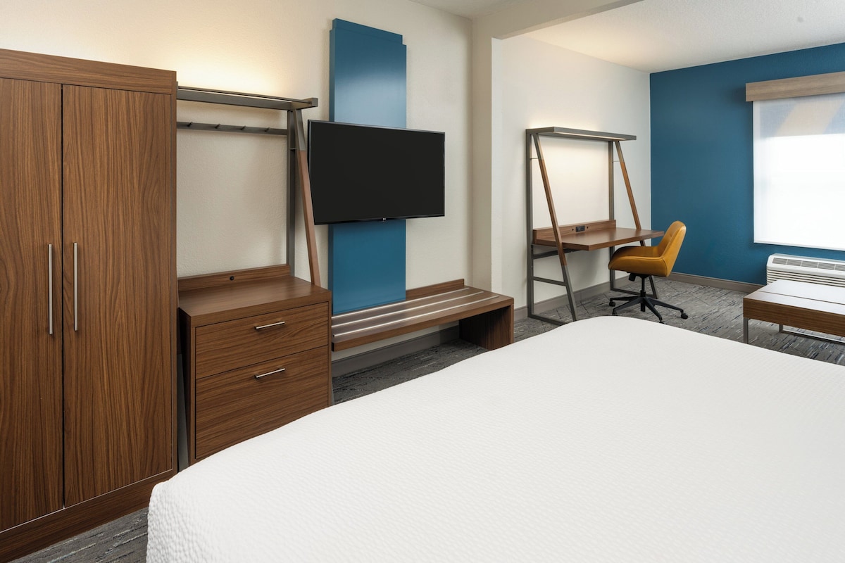 Une chambre d’hôtel avec un grand lit, une armoire en bois, un bureau avec une chaise, un téléviseur fixé au mur et une fenêtre.