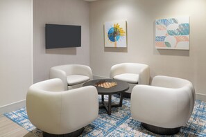 Une pièce moderne avec quatre chaises blanches disposées autour d’une table centrale, un téléviseur à écran plat accroché au mur et deux tableaux abstraits.