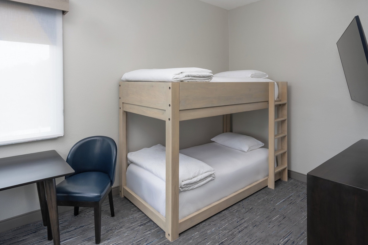 Une chambre avec un lit superposé, un bureau et une chaise, un téléviseur fixé au mur et une fenêtre.