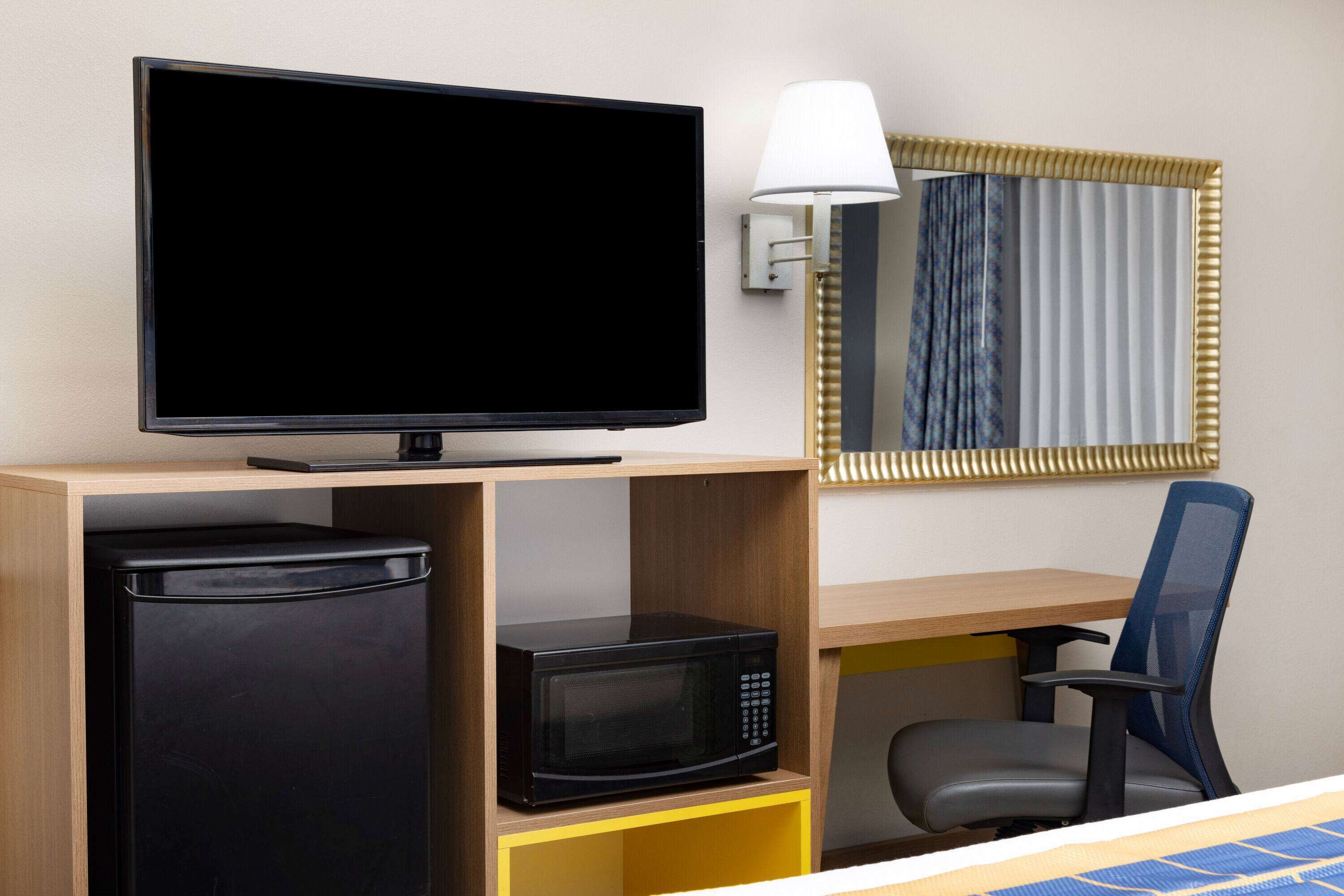 A hotel room with a flat-screen TV, a microwave, a refrigerator, a desk, a chair, and a lamp.