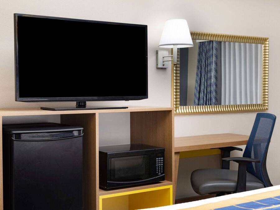 A hotel room with a flat-screen TV, a microwave, a refrigerator, a desk, a chair, and a lamp.