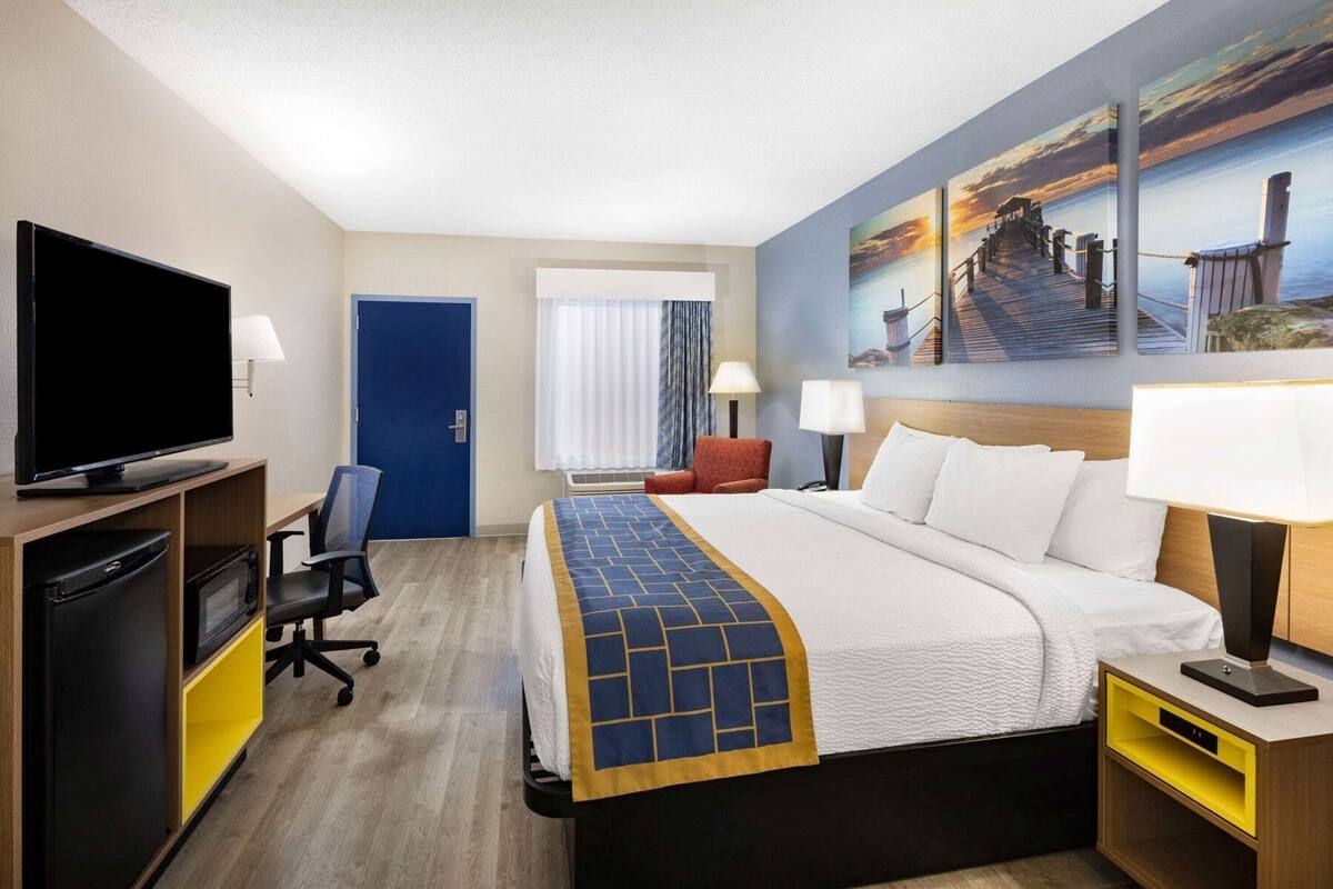 Une chambre d’hôtel comprenant un lit, une télévision, un bureau avec une chaise, un four à micro-ondes, un mini-réfrigérateur, une porte et une fenêtre avec des rideaux.