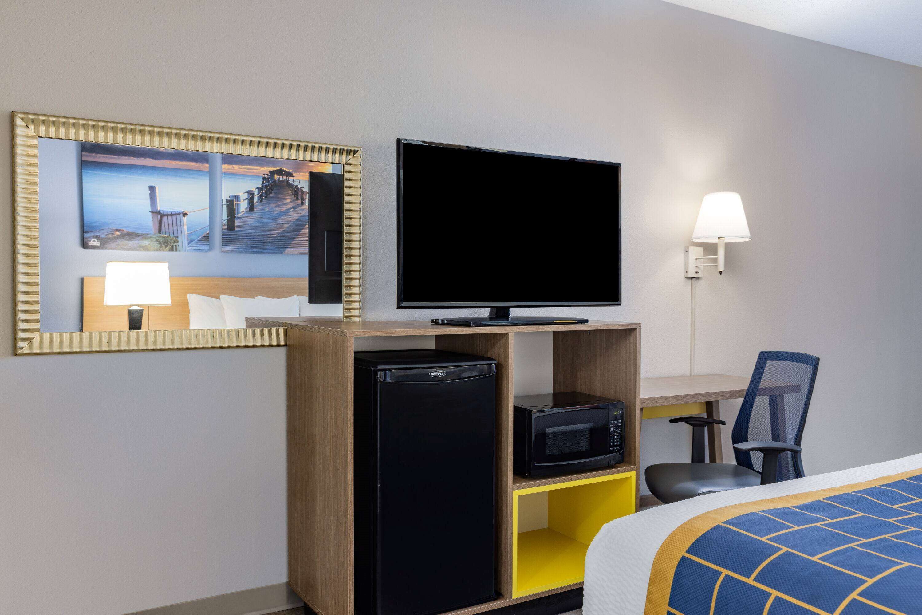 A hotel room with a bed, a television on a wooden shelf, a small refrigerator, a microwave, a desk with a chair, and a painting on the wall.