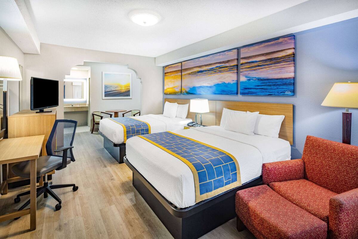 Une chambre d’hôtel avec deux lits, un bureau, une télévision et un tableau représentant un coucher de soleil.