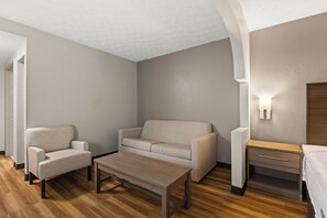 A modern hotel room with a sofa, armchair, wooden coffee table, and a bedside table with a lamp. - Red Roof Inn & Suites Newnan (Newnan)