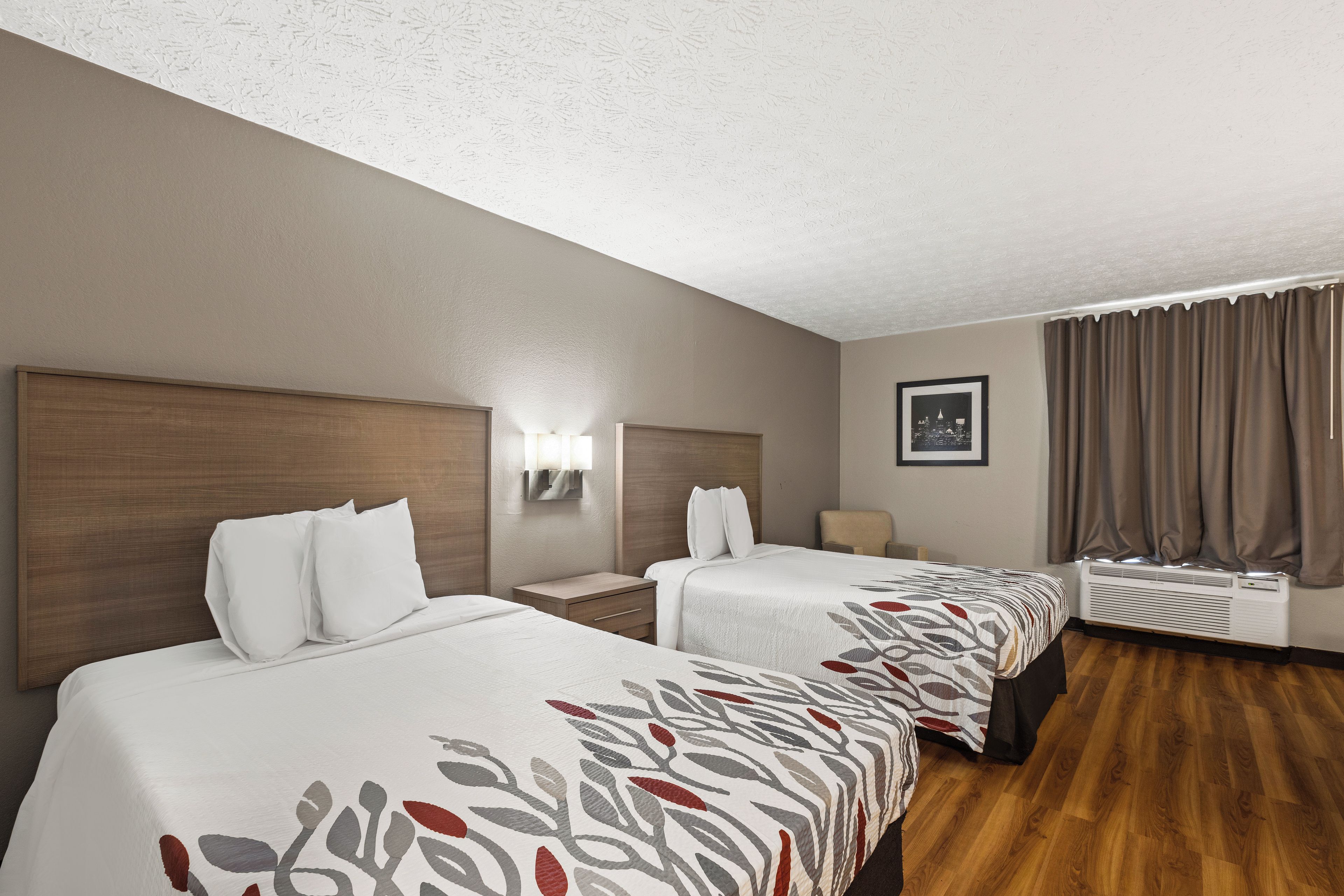 A hotel room with two beds, a nightstand, a wall-mounted lamp, a picture, and a window with curtains.