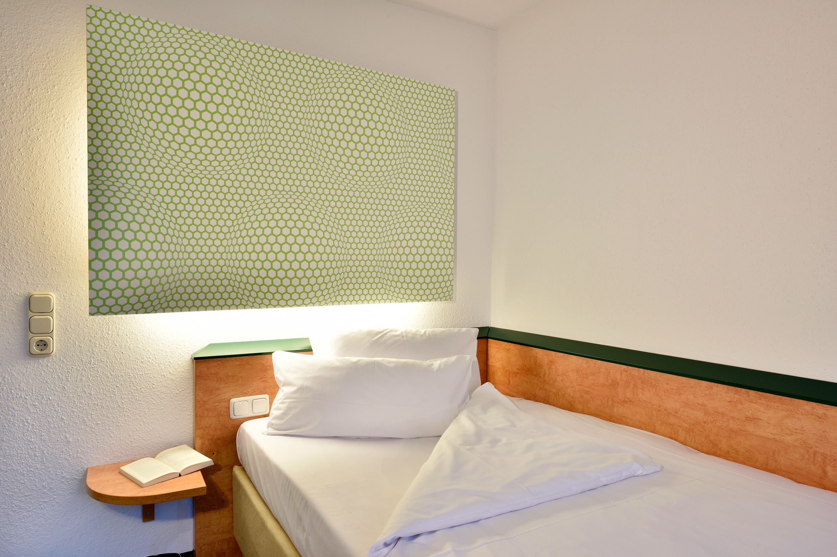 A hotel room with a neatly made bed, a bedside table with a book, and a wall-mounted light fixture.