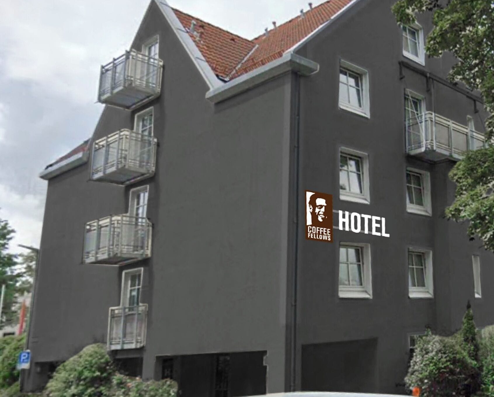 Um edifício de vários andares com fachada cinzenta e telhado de telhas vermelhas. Possui várias varandas e uma placa indicando que é um hotel.