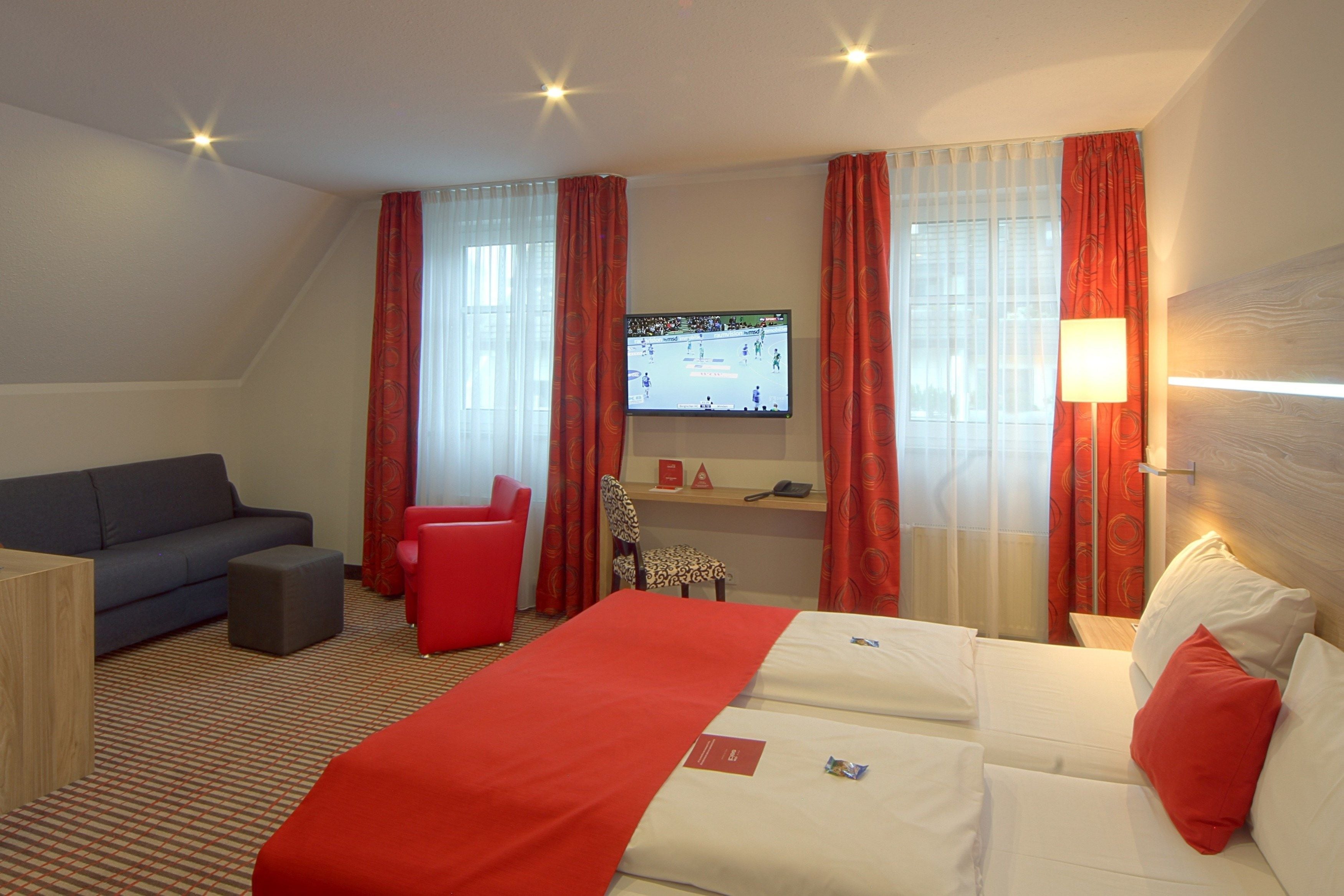 A hotel room with a bed, a sofa, a red chair, a TV, and a desk.