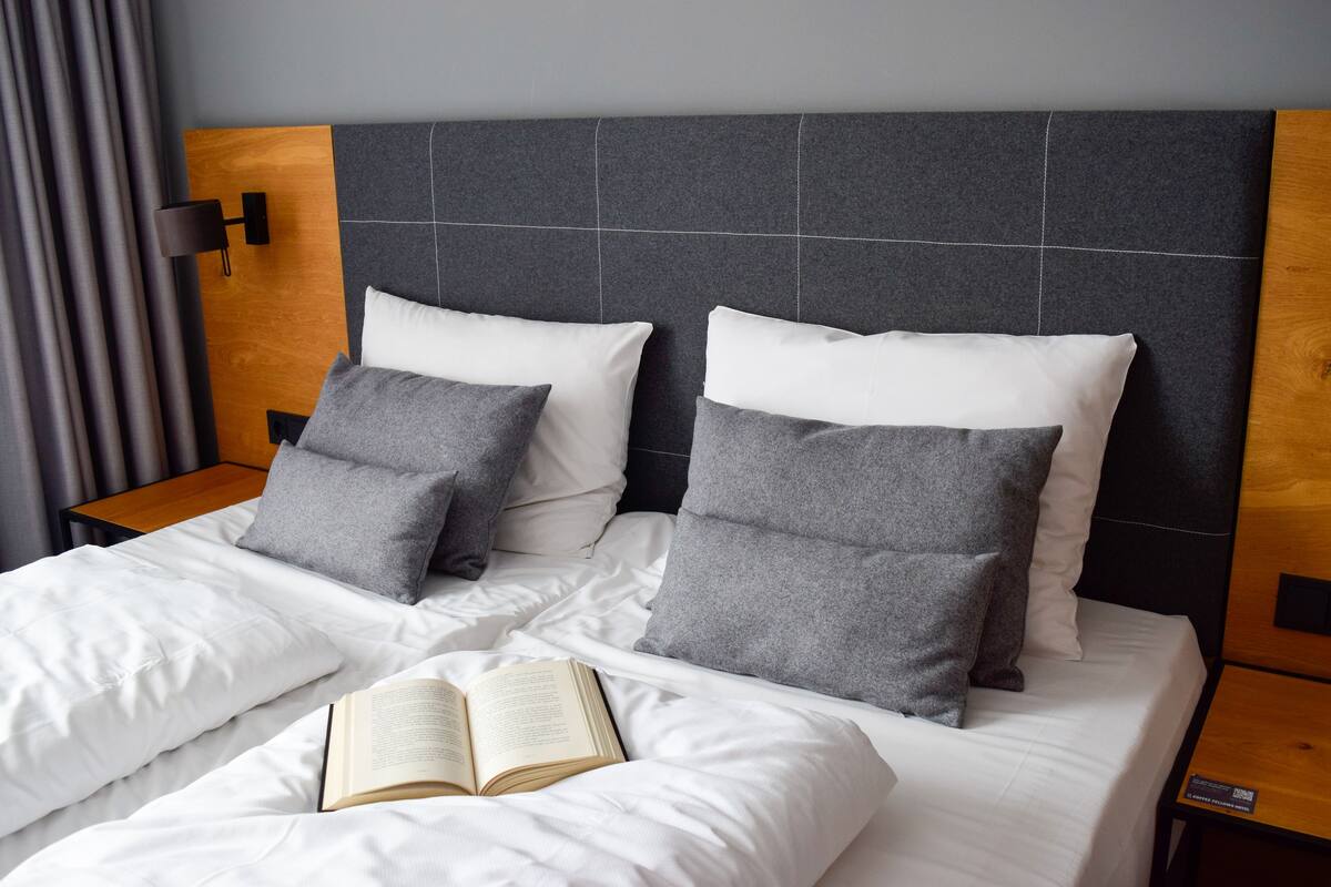 Ein ordentlich bezogenes Bett mit weißer und grauer Bettwäsche, ein Buch auf dem Bett und ein moderner Kopfteil.