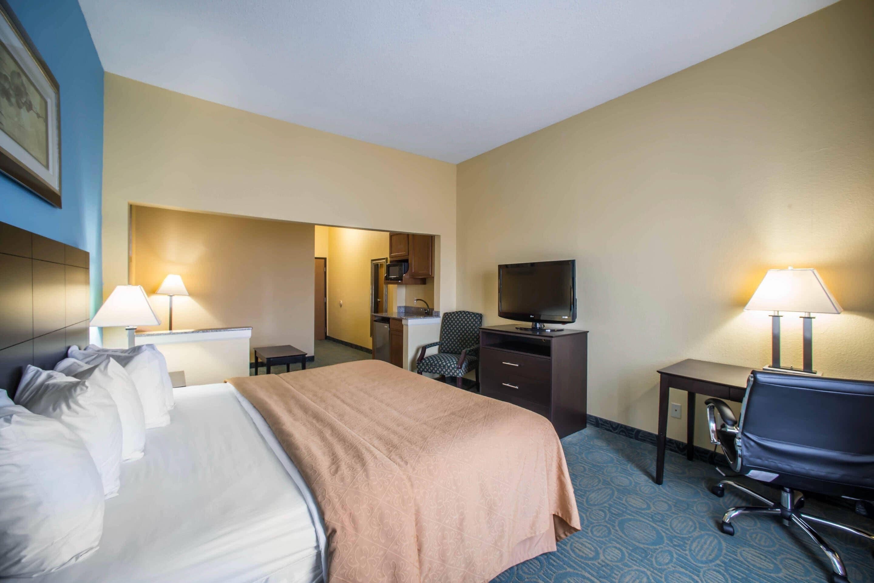 A hotel room with a bed, desk, chair, television, and a kitchenette.