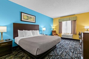 Ein Hotelzimmer mit einem großen Bett, zwei Nachttischen, einem Sessel, einem Schreibtisch, einem Fenster mit Vorhängen und einer Wand-Klimaanlage.