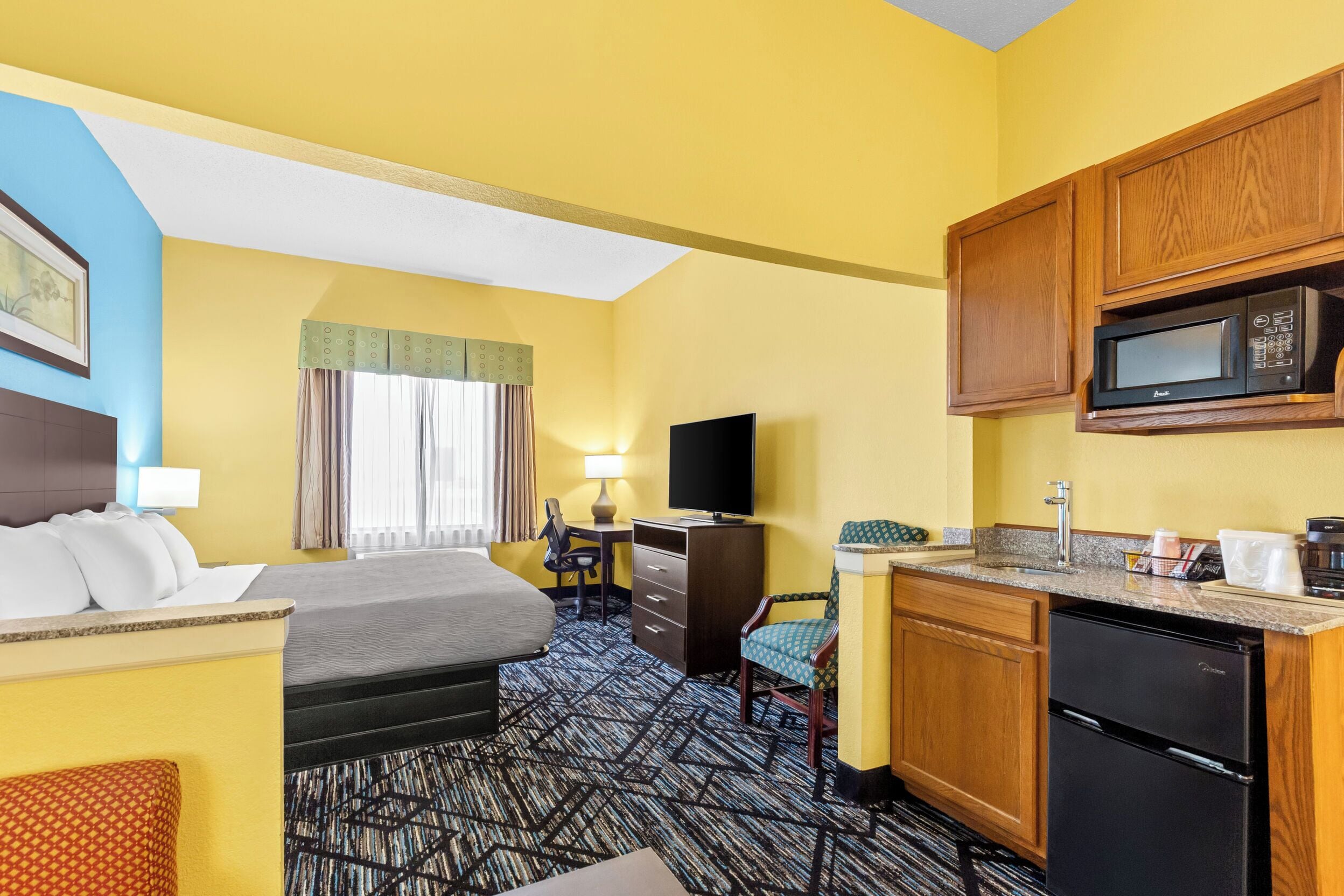 A hotel room with a bed, desk, chair, kitchenette, and a television.