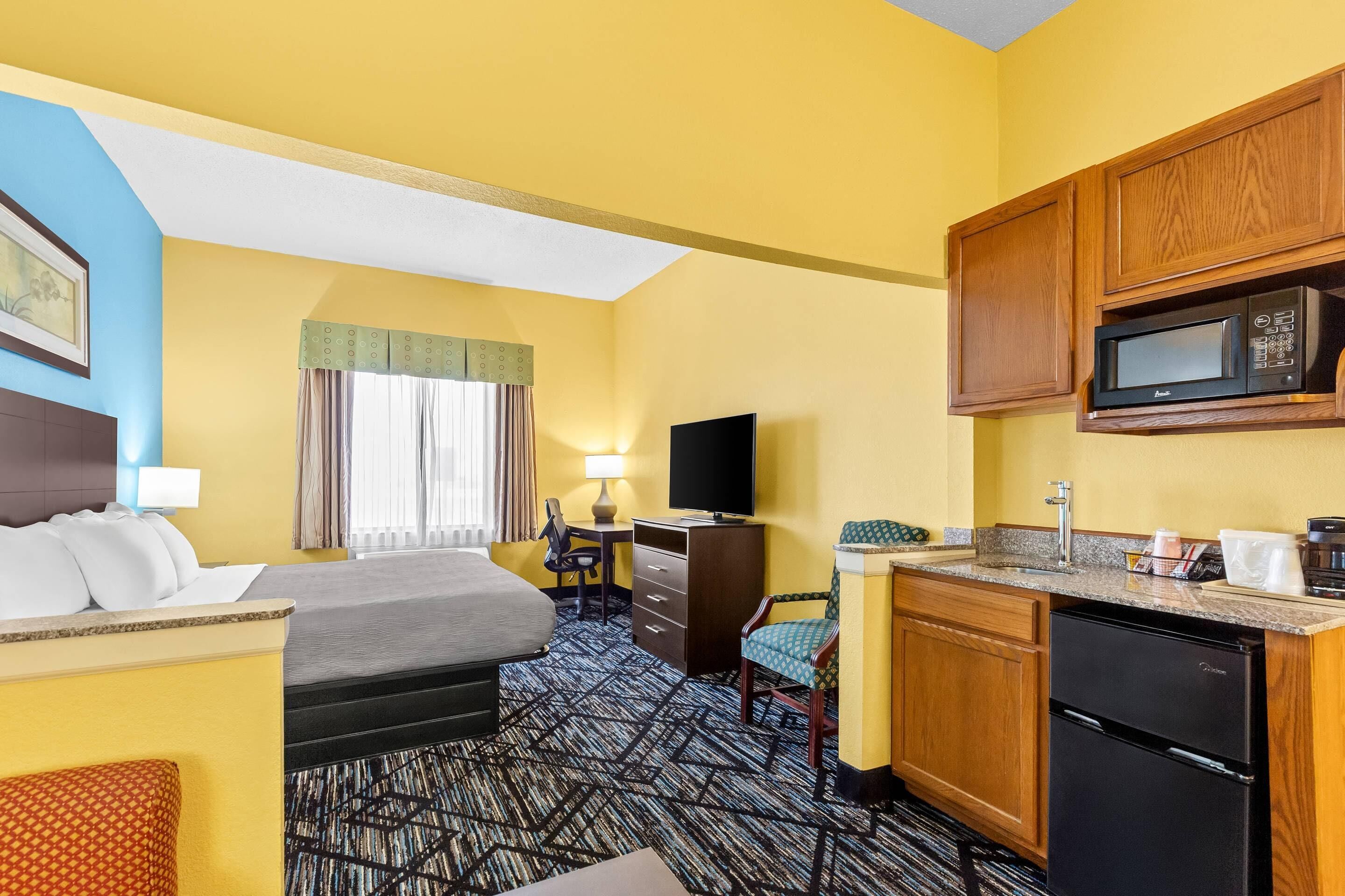 A hotel room with a bed, desk, chair, kitchenette, and a television.