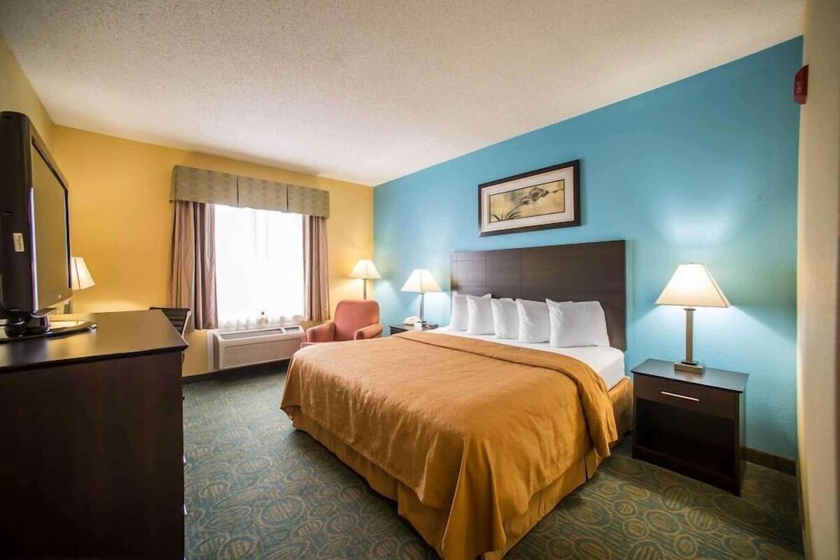Habitación de hotel con una cama grande, dos mesitas de noche con lámparas, un escritorio con computadora y una ventana con cortinas.