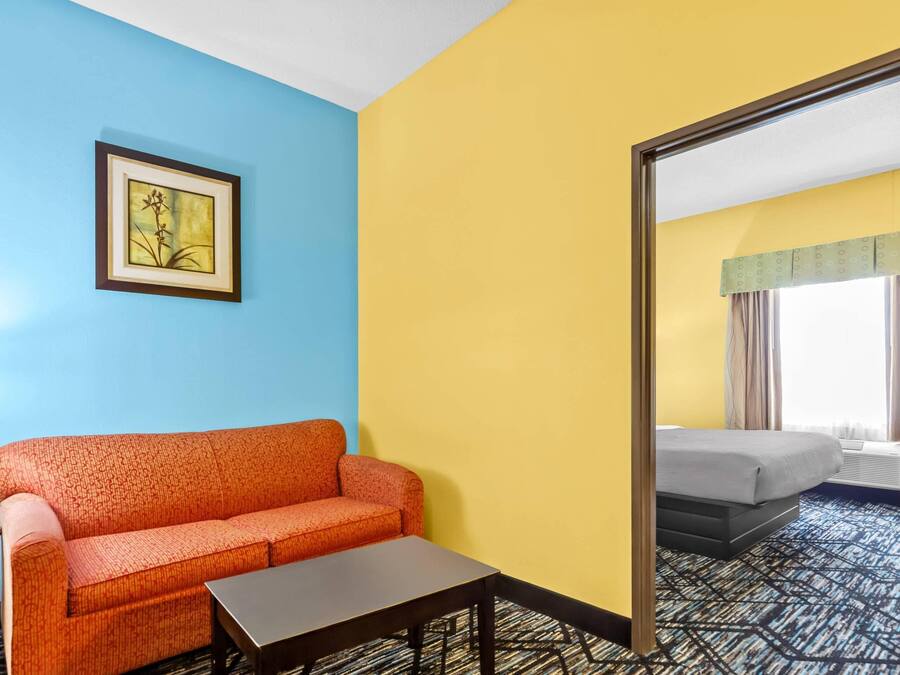 A hotel room with a blue wall, a yellow wall, a framed picture, a red sofa, a small table, and a bed with a window view.
