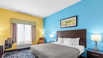 Quarto de hotel com cama, duas luminárias de cabeceira, uma cadeira, uma escrivaninha com computador e uma janela com cortinas.