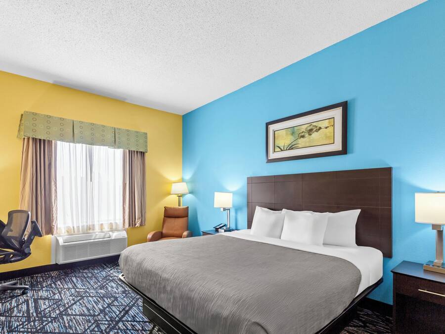 A hotel room with a bed, two bedside lamps, a chair, a desk with a computer, and a window with curtains.