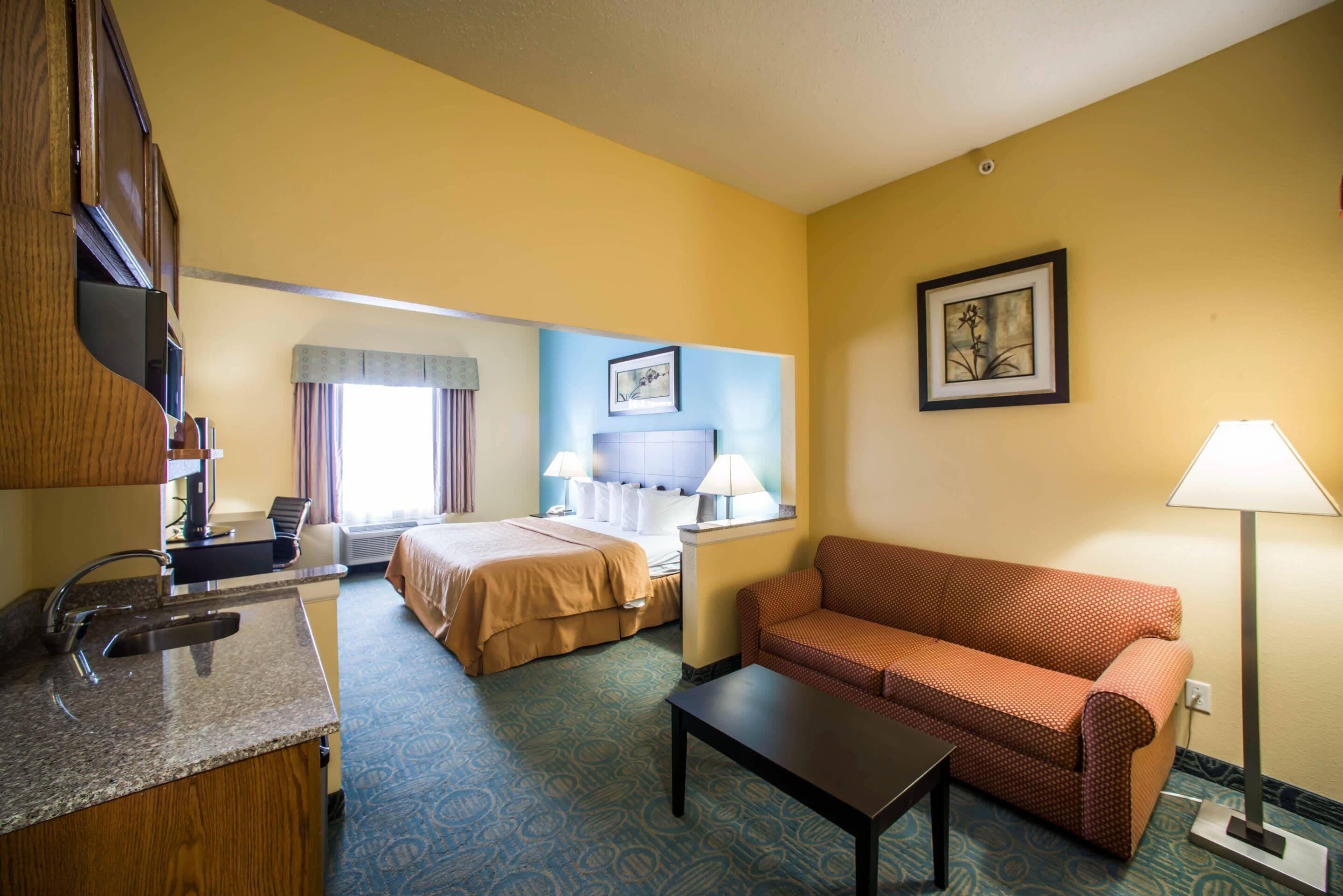 A hotel room with a bed, a sofa, a desk with a lamp, and a painting on the wall.