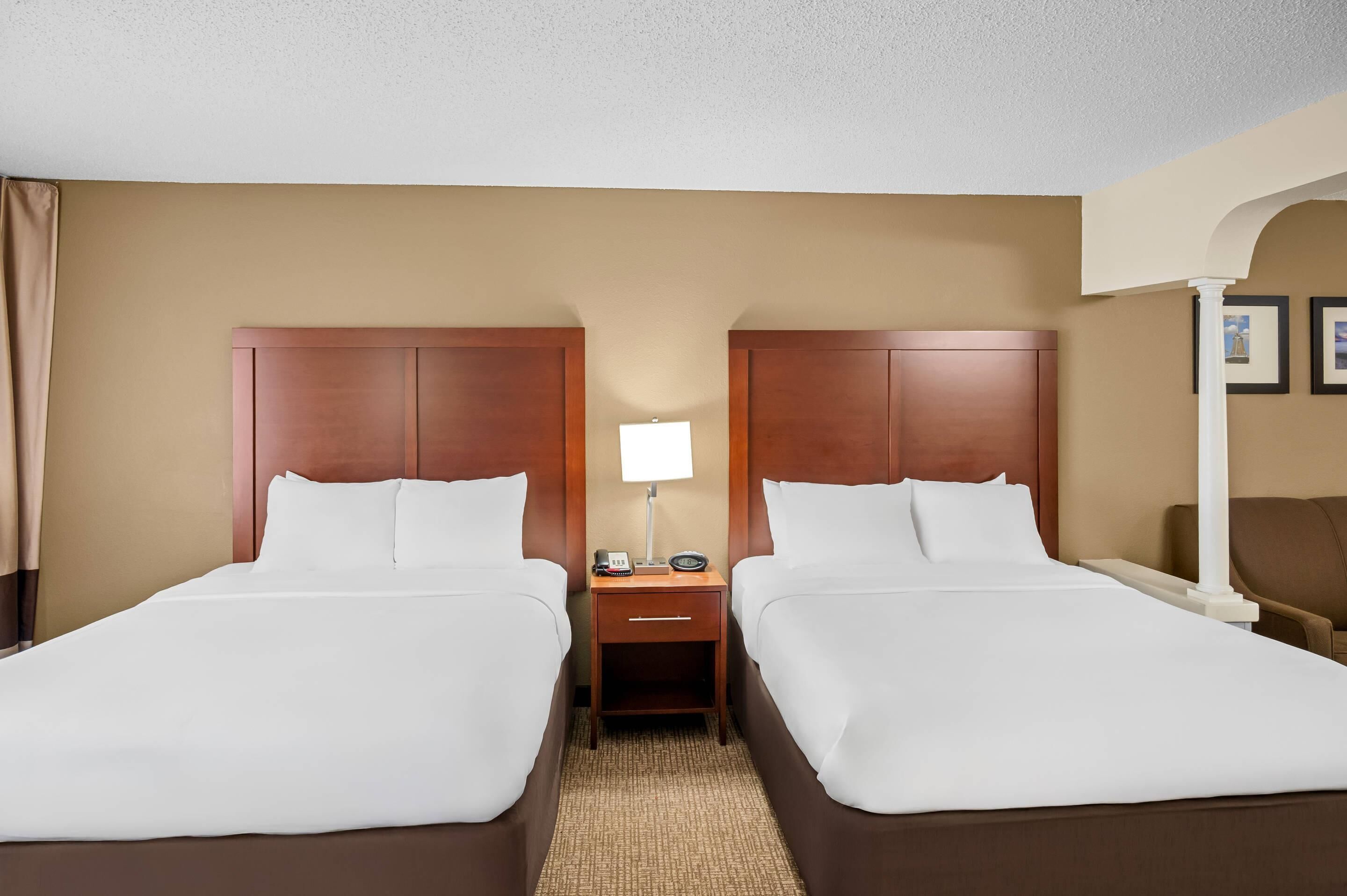 A hotel room with two beds, a nightstand with a lamp, and a chair.