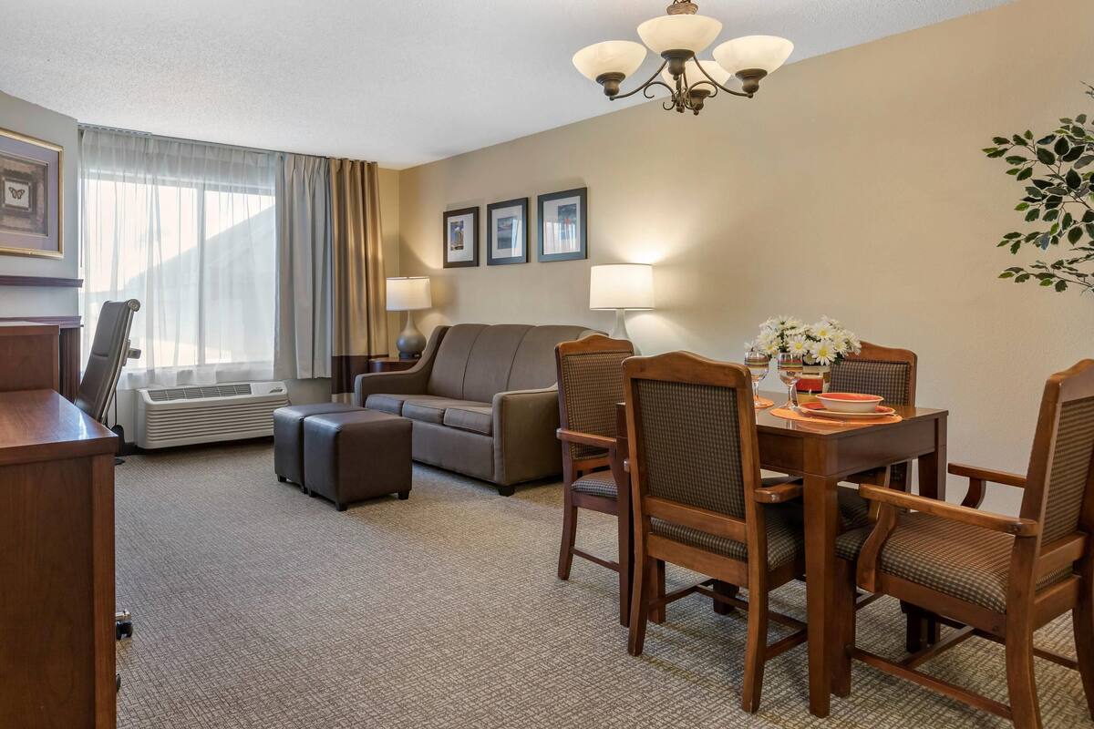 Una habitación de hotel con sala de estar, una mesa de comedor con sillas, un sofá, una mesa auxiliar con un jarrón de flores y un lámpara en el techo.