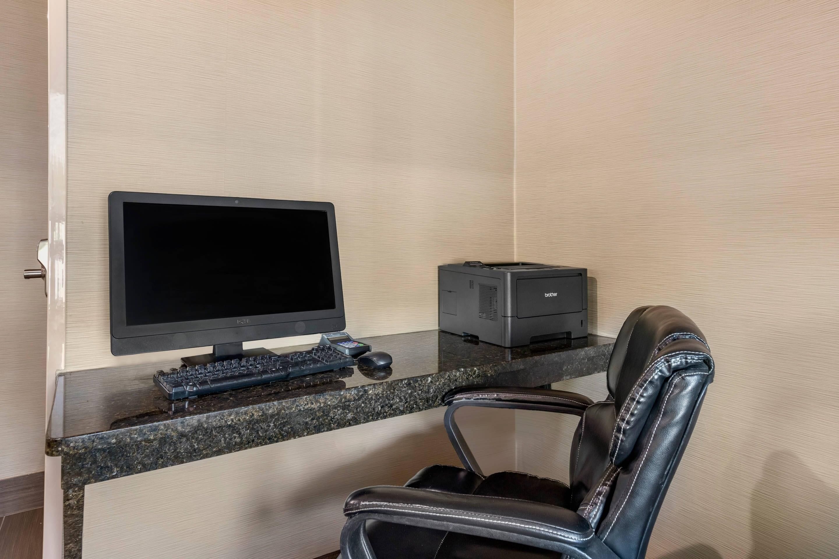 A hotel room with a desk, a computer monitor, a printer, and a leather chair.