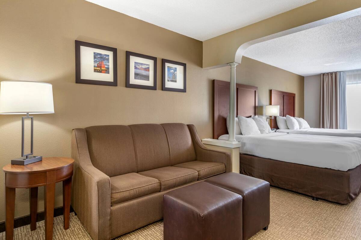 Una habitación de hotel con un sofá marrón, una mesita con una lámpara, una cama grande y dos cuadros en la pared.