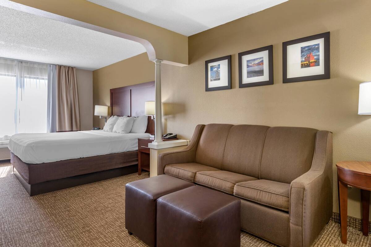 Habitación de hotel con una cama, un sofá, un reposapiés, una mesita de noche, una lámpara y tres cuadros en la pared.