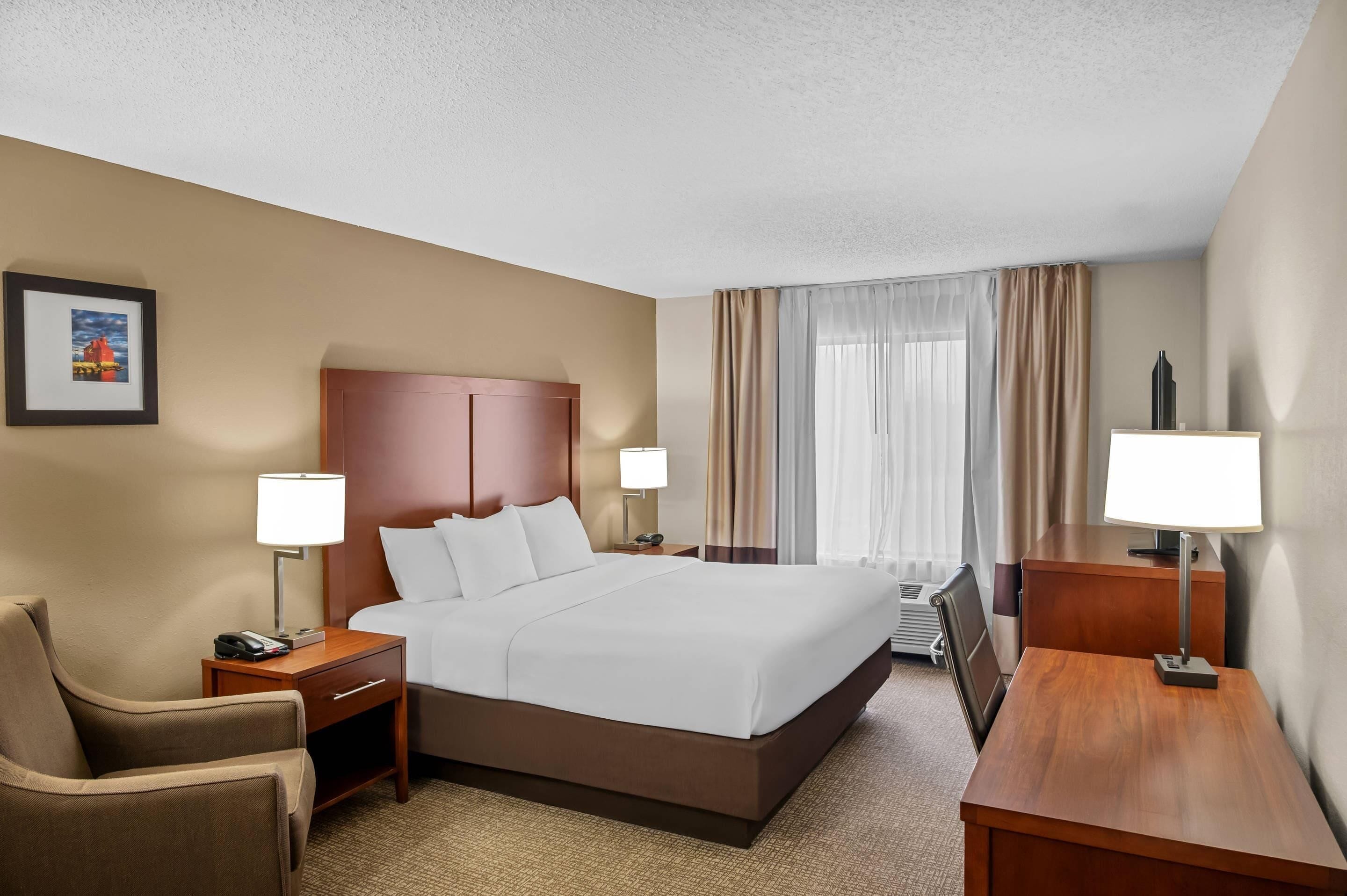 A hotel room with a large bed, two bedside tables with lamps, a chair, a desk, and a picture on the wall.