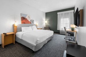 A hotel room with a bed, bedside tables, a desk with a chair, a lamp, a painting, and a window with curtains. - Spark by Hilton Grain Valley Kansas City (Grain Valley)