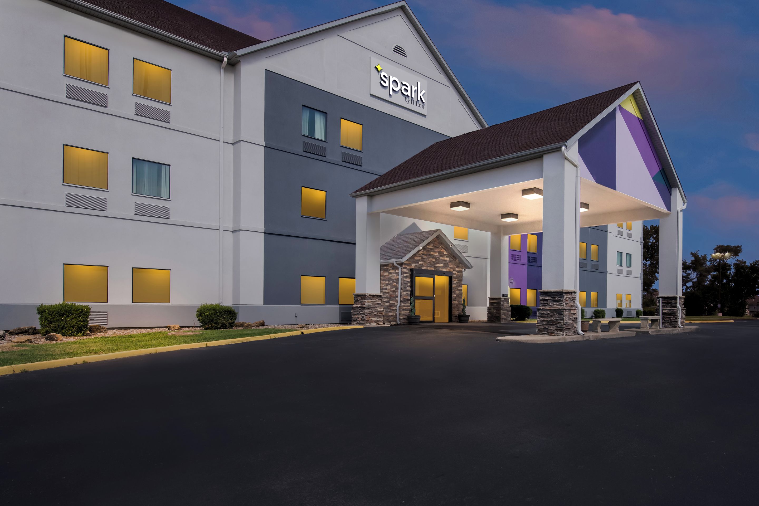 A hotel with a modern design, featuring a prominent 'spark by Hilton' sign, a covered entrance, and multiple windows with illuminated interiors.