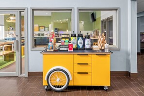 A yellow cart with beverage dispensers and a wheel, located in a room with large windows and a view of a kitchen area. - Spark by Hilton Grain Valley Kansas City (Grain Valley)