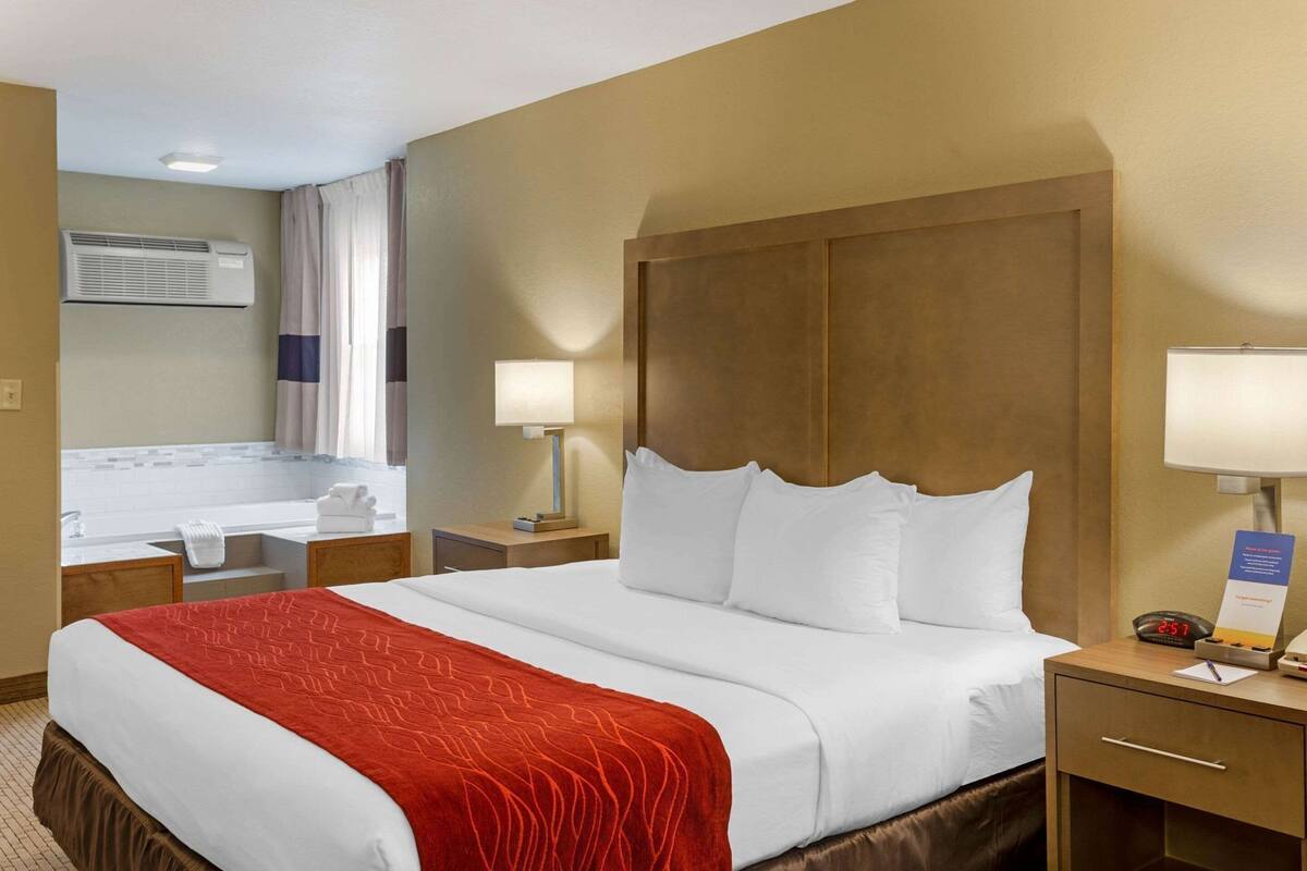 Habitación de hotel con cama, mesita de noche con lámpara, escritorio con lámpara, baño con lavamanos y espejo, y ventana con cortinas.
